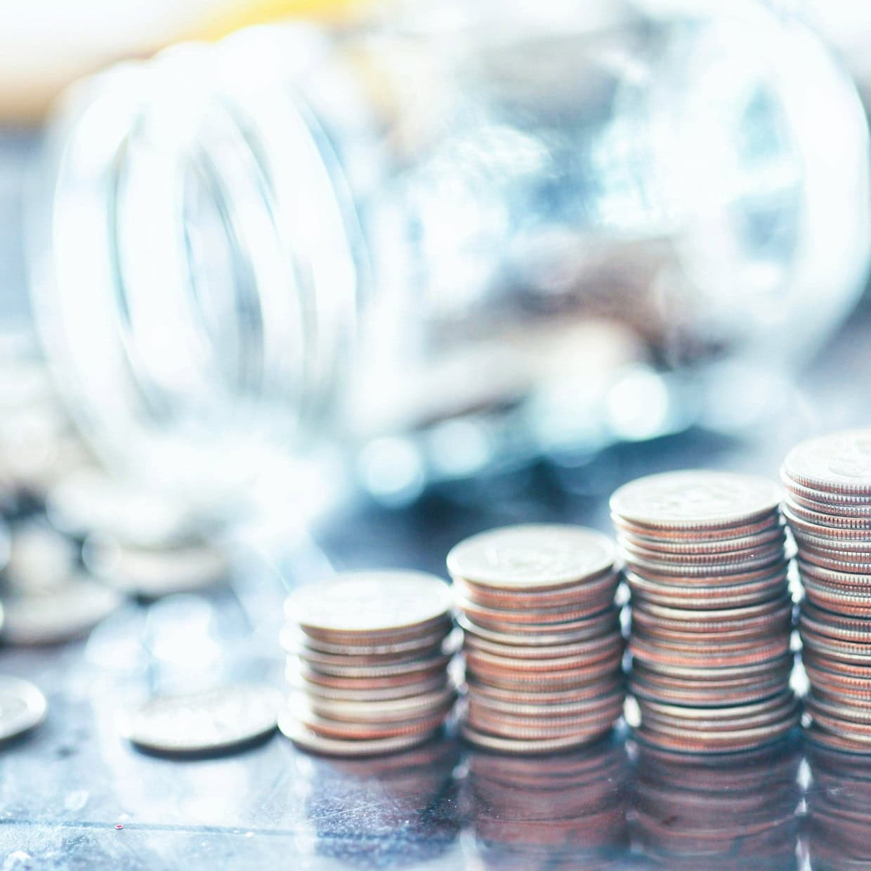 Stacks of coins arranged in ascending order beside a glass jar on a reflective surface.