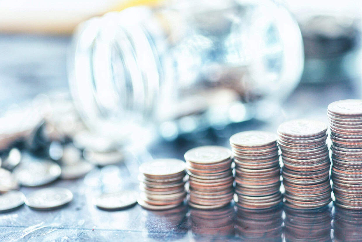 Stacks of coins arranged in ascending order beside a glass jar on a reflective surface.