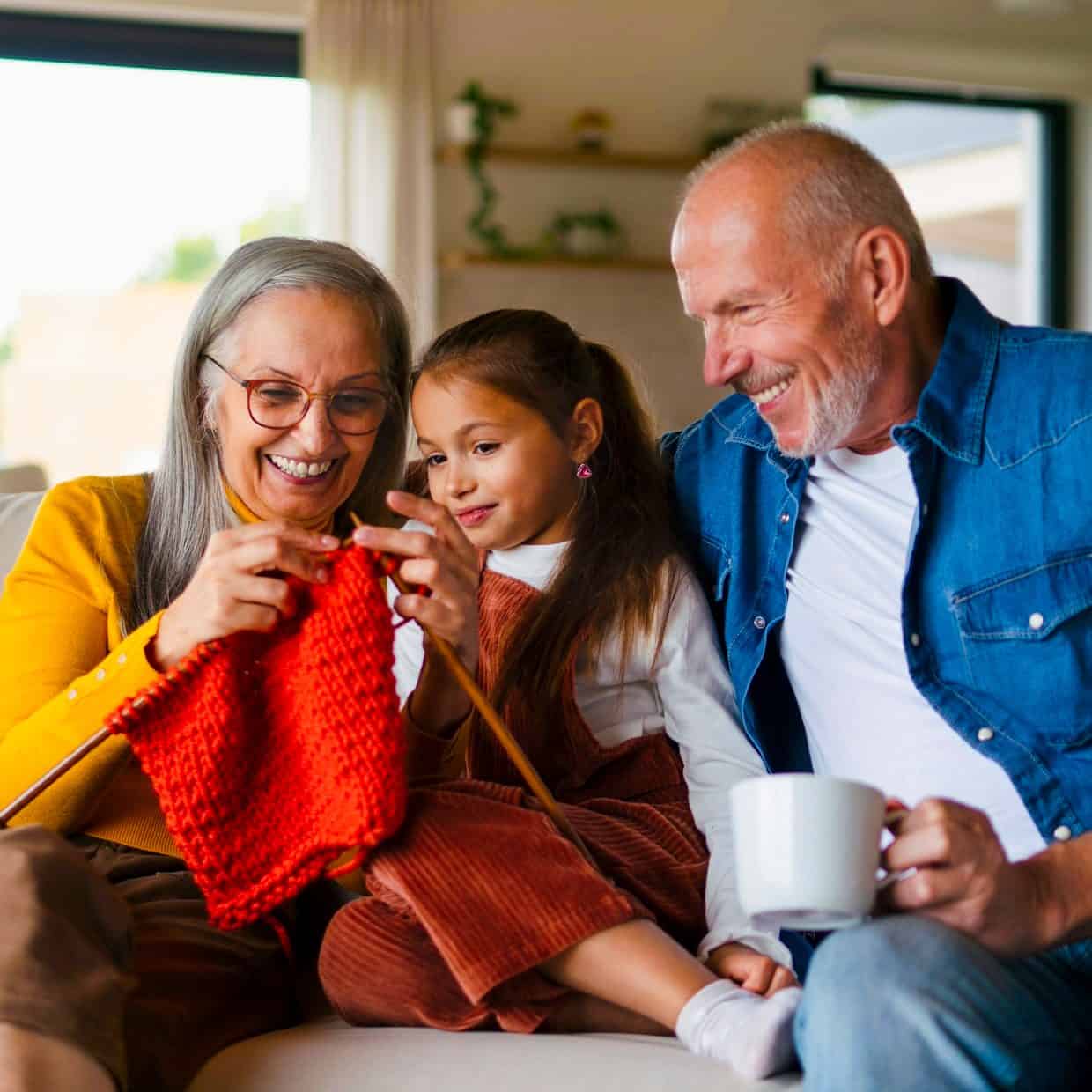 Smiling grandparents sitting on a sofa with their granddaughter, teaching her how to knit with bright orange yarn.
