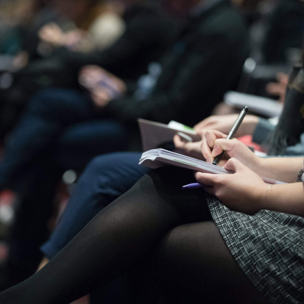 Close-up of a person seated in an audience writing notes in a notebook during a presentation, with other attendees blurred in the background.