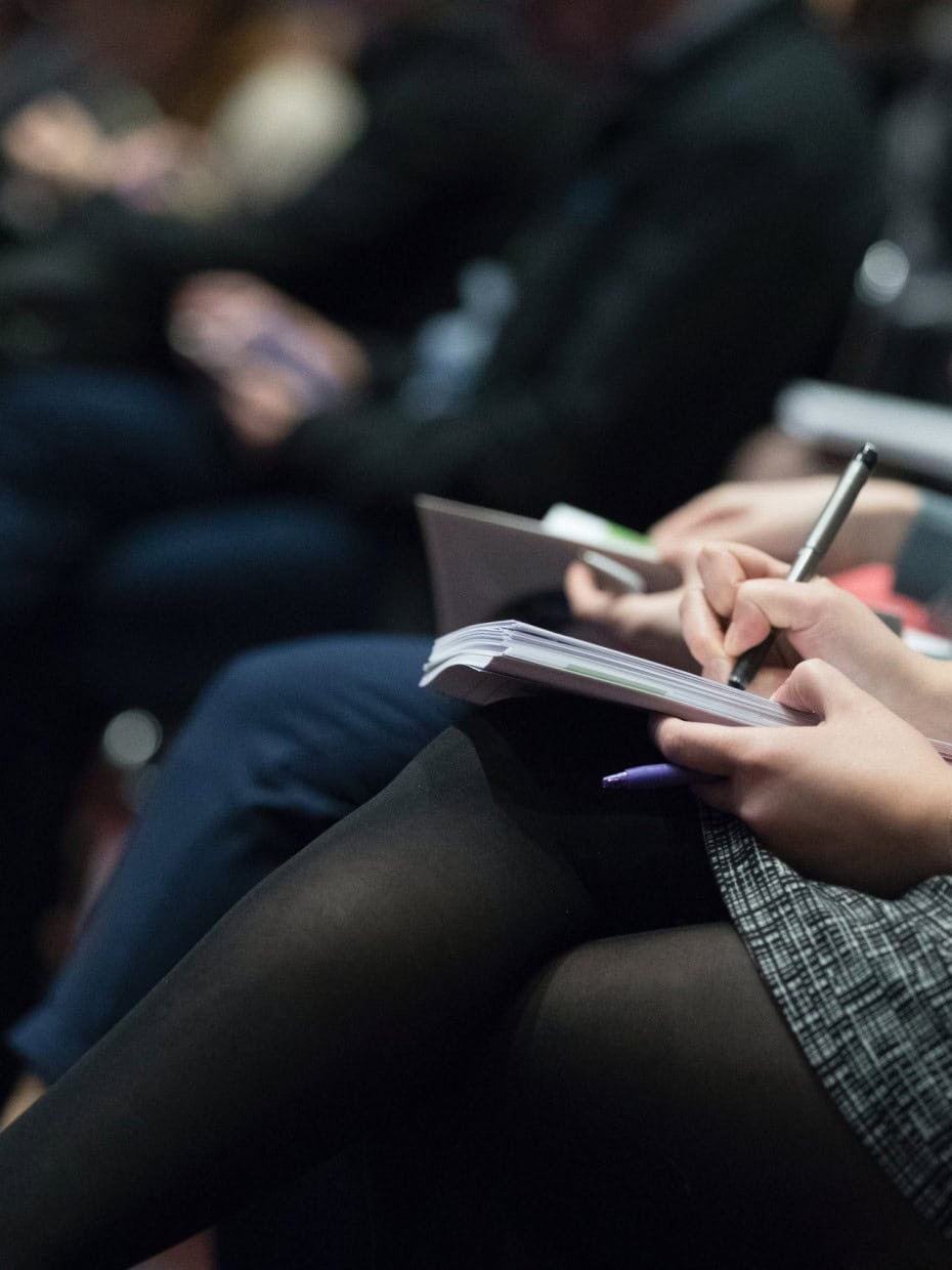 Close-up of a person seated in an audience writing notes in a notebook during a presentation, with other attendees blurred in the background.