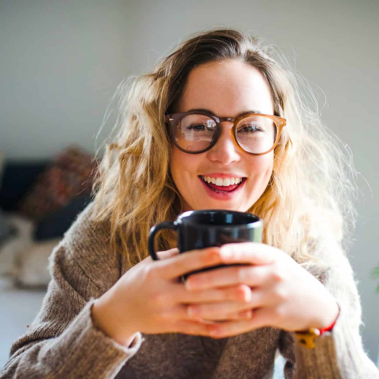 Smiling woman with wavy hair and glasses holding a black mug, sitting indoors in a cosy setting.