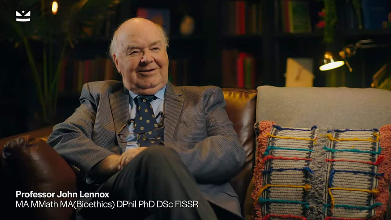 Man in a suit sitting on a leather sofa in a warmly lit room with bookshelves in the background, with on-screen text identifying him as Professor John Lennox.