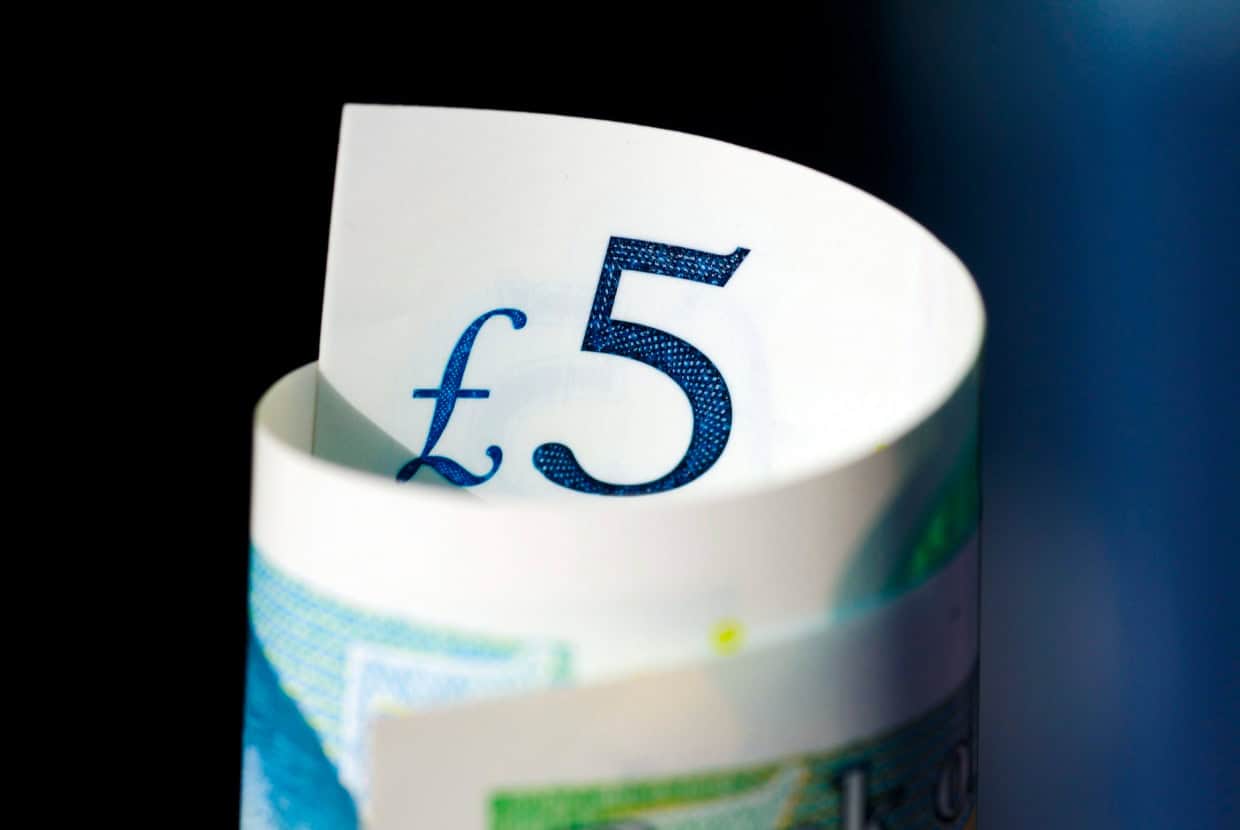 Close-up of a UK five-pound note showing the blue £5 symbol against a dark background.